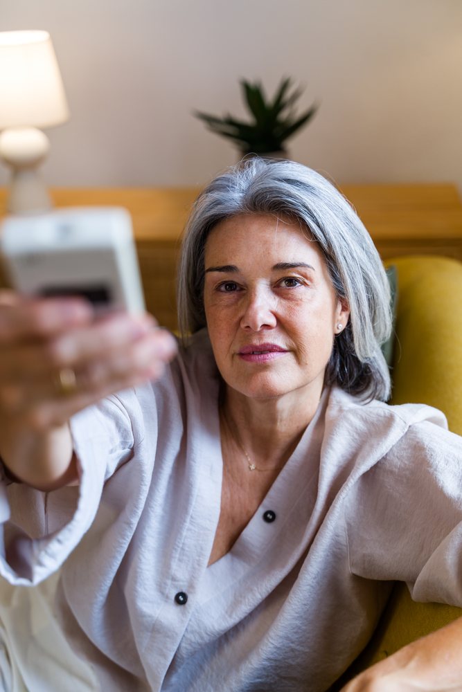 Mature woman with gray hair sitting on a sofa in a living room holding a remote control near a lamp and potted plant, Spain.