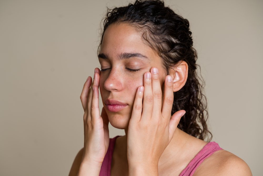Woman with curly hair applies moisturizer to her face wearing a pink tank top against a neutral background during a beauty shoot, Spain.