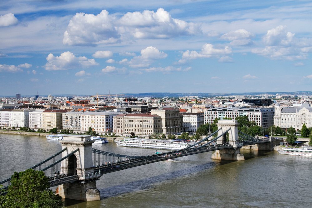 Chain Bridge spans the River Danube with Budapest cityscape and boats under blue sky and clouds, Hungary.