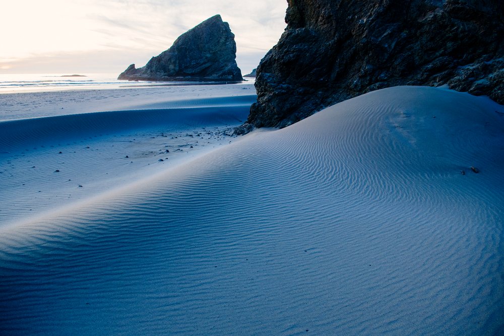 Sand dunes with rippled patterns and rock formations on Meyers Creek beach at sunset with ocean waves approaching the sandy shore, Oregon, USA.