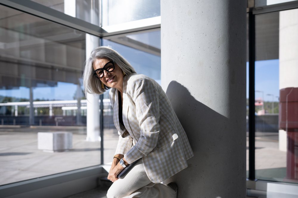 Businesswoman with gray hair wearing glasses and a plaid blazer leans smiling inside a modern glass building, Spain, Madrid.