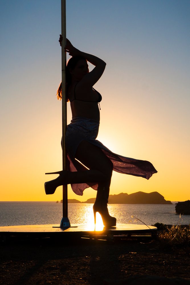 Silhouette of a woman holding a pole dance pole on a platform at sunset by the ocean with islands, Ibiza, Spain.