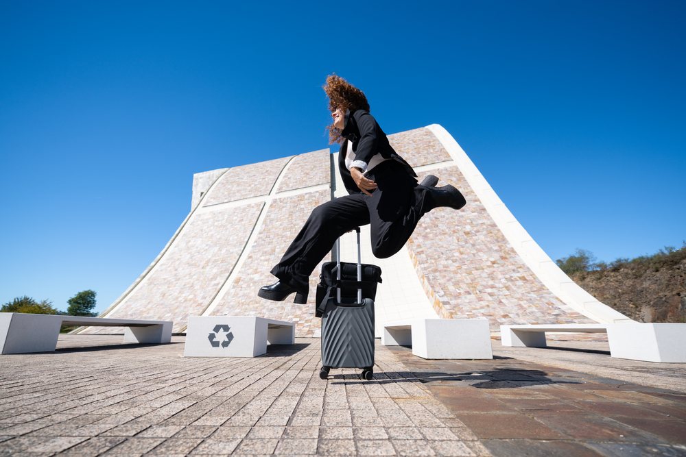 Businesswoman jumping with a wheeled suitcase in an urban plaza with modern curved architecture and blue sky in Santiago de Compostela, traveling executive, Spain, Antiago De Compostela.