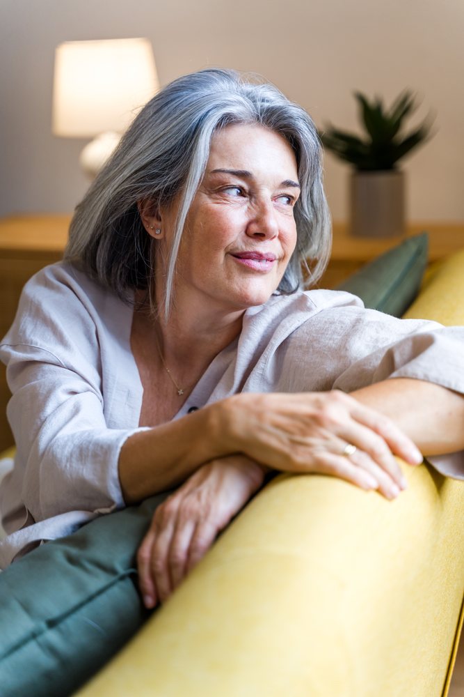 Mature woman with gray hair seated on a yellow sofa indoors, relaxing with a lamp and green houseplant in the background, Spain.