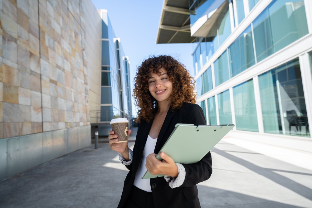 Businesswoman holding a coffee cup and clipboard standing outside a modern office building, traveling executive, Spain.