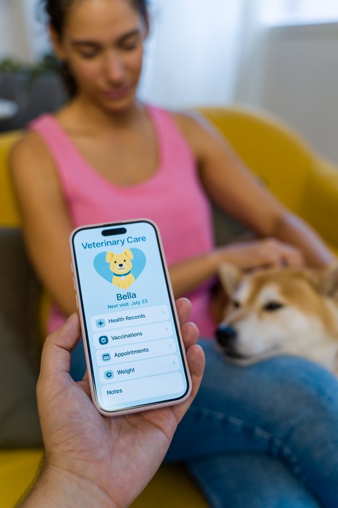 A person holds a smartphone displaying a veterinary care application while a woman pets her dog on a yellow couch, Spain.