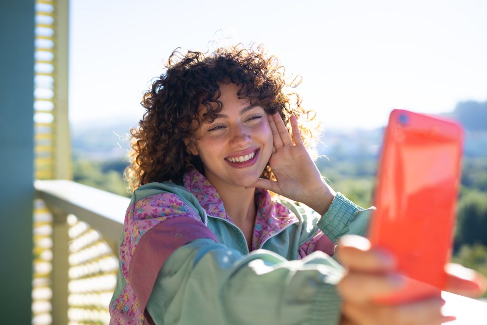 Smiling woman with curly hair takes a selfie outdoors in daylight wearing a casual jacket, Spain.
