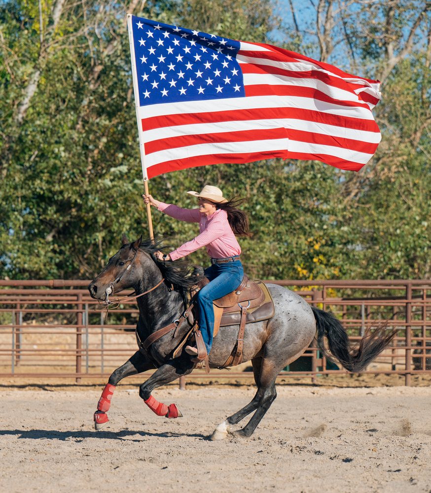 Woman wearing a Western hat gallops a horse in an outdoor sandy arena while holding a large American flag, Southwest USA.