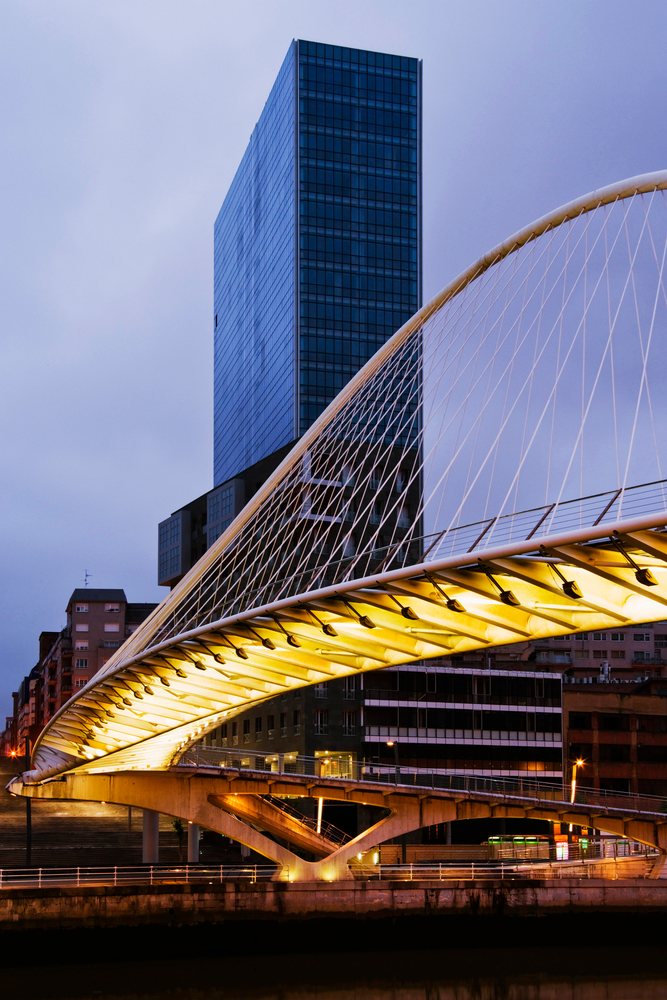 Illuminated modern pedestrian suspension bridge over a river with city buildings at dusk, Bilbao, Spain.