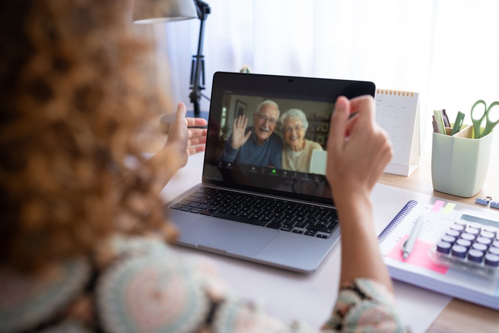 Woman with curly hair waving during a video call with an elderly couple on a laptop at a home office desk, Spain.