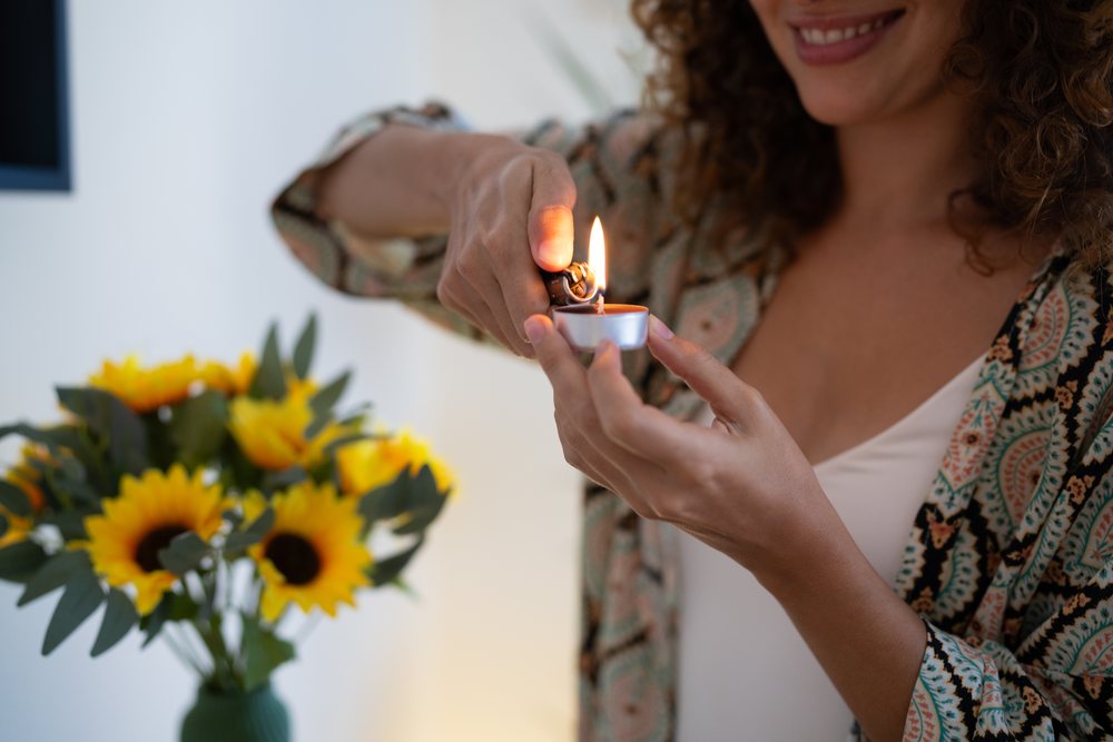 Woman lighting a tealight candle near a green ceramic vase of bright yellow sunflowers in an indoor home setting, Spain.