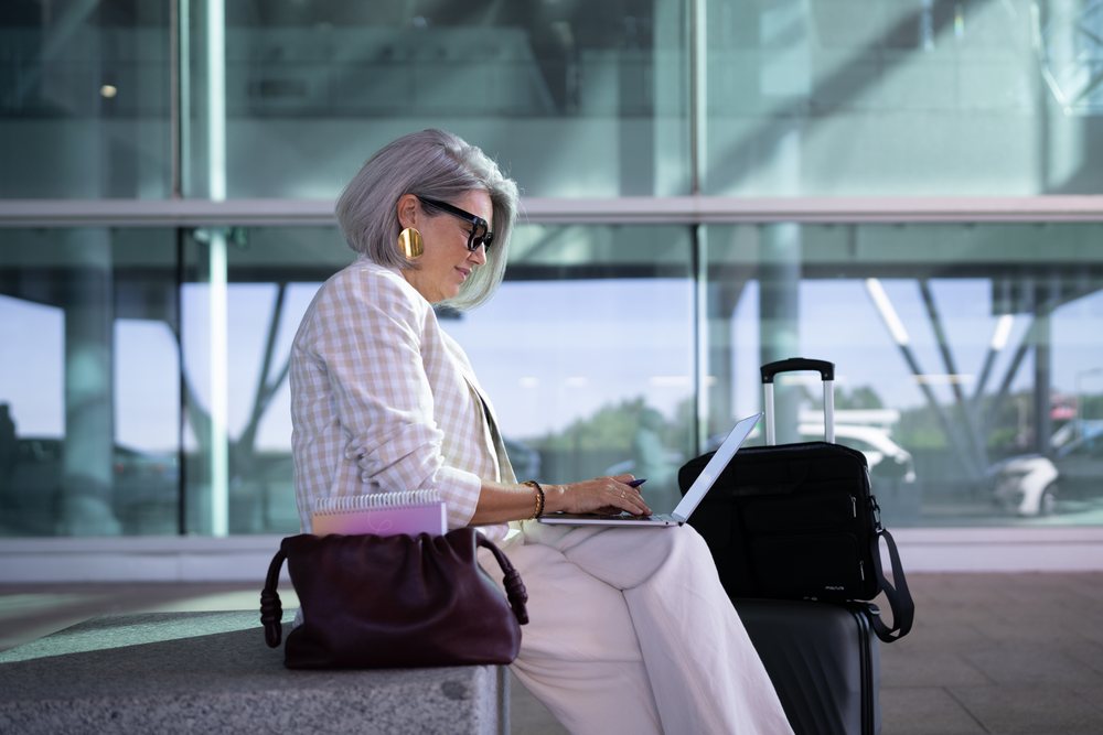 Businesswoman with gray hair working on laptop while seated outdoors with rolling suitcase and handbag at airport, Spain, Madrid.