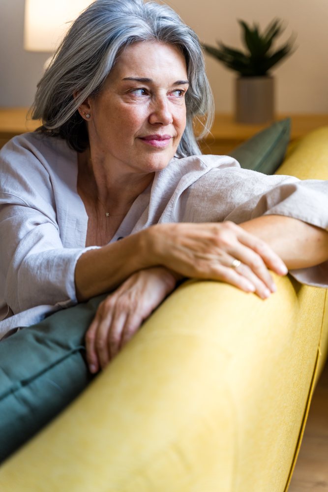Mature woman with gray hair sitting thoughtfully on a yellow sofa in a cozy indoor home setting with a potted plant nearby, Spain.