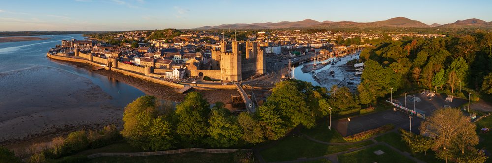 Aerial view showing Caernarfon Castle and town at Sunset with a river, boats, parks, and rolling hills, Landscape, Gwynedd, Wales.