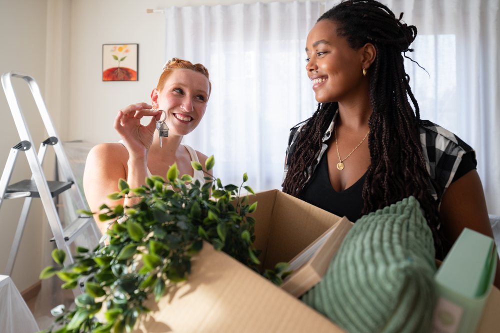 Two women smiling and unpacking a box with houseplant and home decor items as they hold keys during a move, moving, Spain.