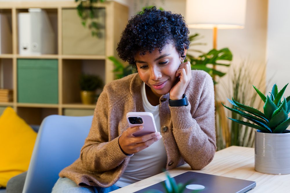 Young woman wearing a brown cardigan sits at a table indoors using a smartphone, with a closed laptop and plants nearby, Spain.