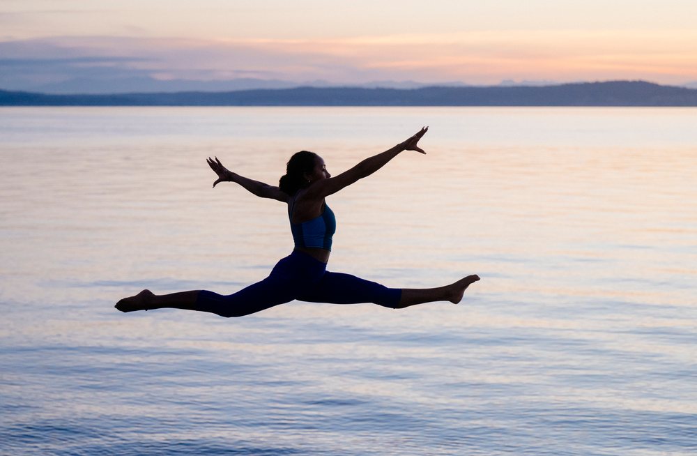 Woman performing a jumping split yoga pose over a calm lake at sunset, USA.