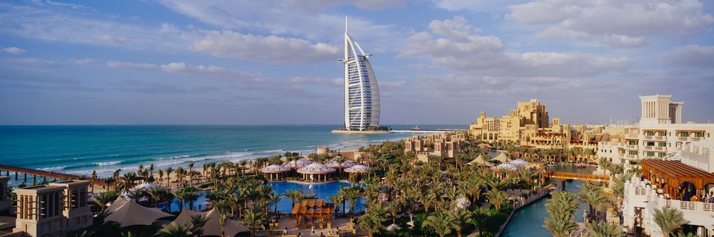 View of the Burj Al Arab hotel and coastal resort along the Arabian Gulf under a partly cloudy sky.