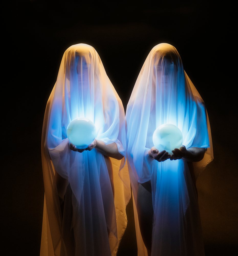 Two people covered in white translucent veils each hold glowing blue orbs against a dark background, ghosts, USA.