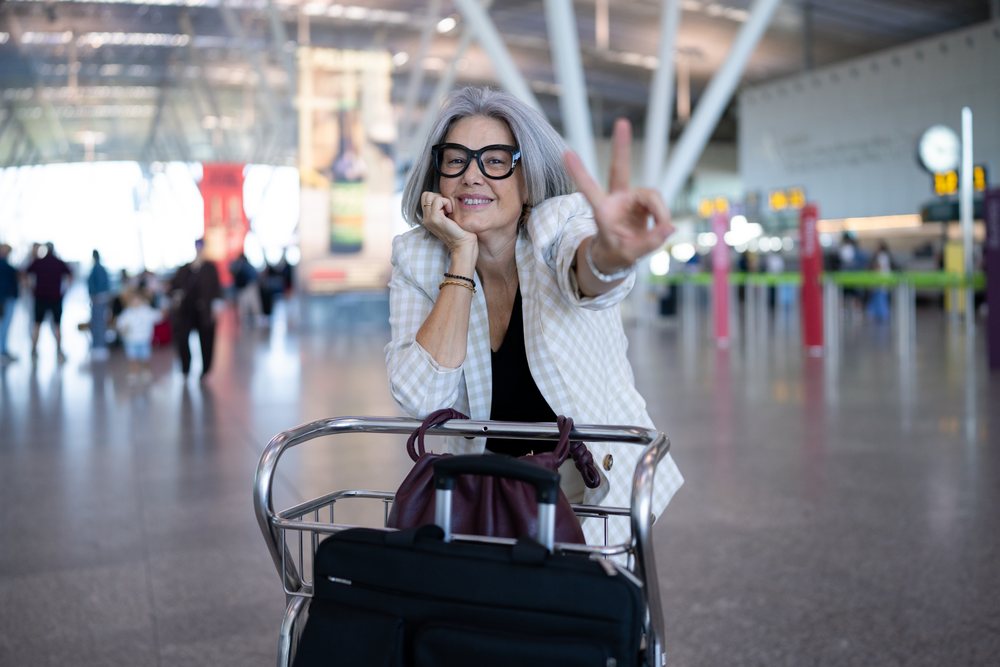 Woman with silver hair and glasses making a peace sign while waiting with luggage trolley at an airport terminal, businesswoman, traveler, Spain, Madrid.