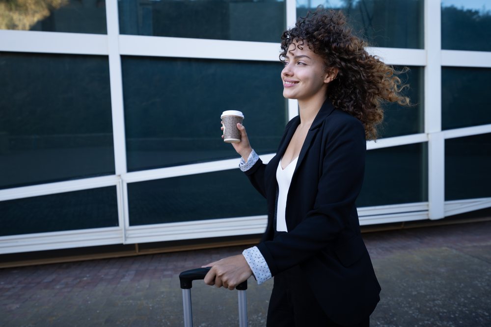 Businesswoman walking outside a modern building with a rolling suitcase and coffee cup, business trip, Spain, Santiago De Compostela.