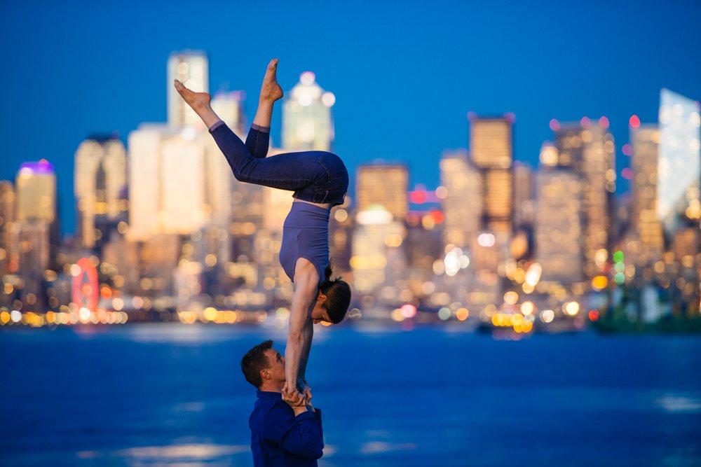 Two people perform partner acrobatics yoga at Seattle's urban waterfront during twilight with city skyline and lights in the background, USA.