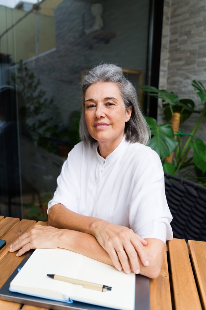 Middle-aged woman sitting at a wooden table with a notebook and pen in a modern indoor space with plants, Spain.