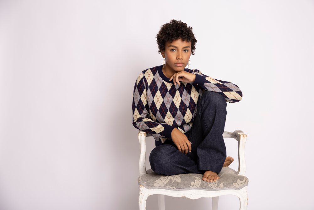 Young woman wearing an argyle sweater sitting barefoot on a white vintage chair in a studio with a white background, fashion, young culture.