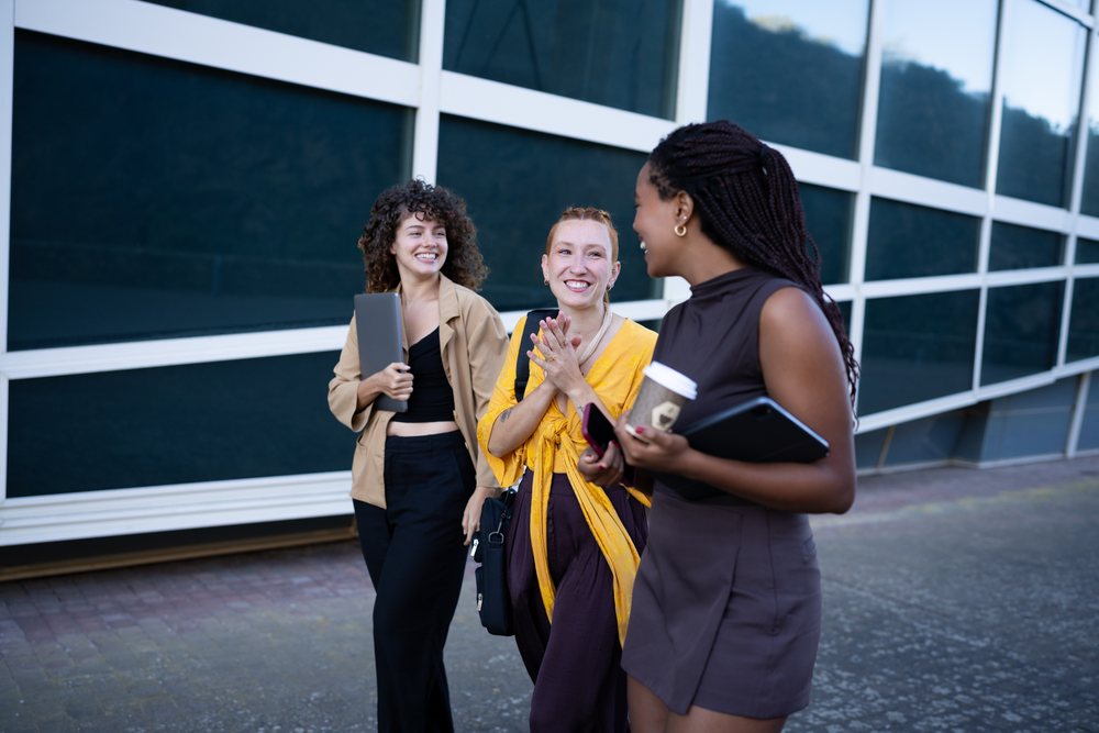 Three young women walk and chat happily near a glass building on an urban sidewalk in daytime, Spain.