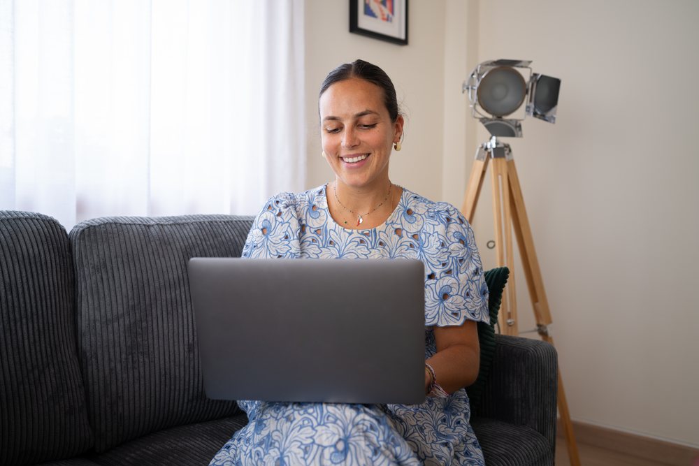 Woman in a blue floral dress smiling while working on a laptop seated on a grey sofa indoors with a modern floor lamp visible behind her, telecommuting, Spain.