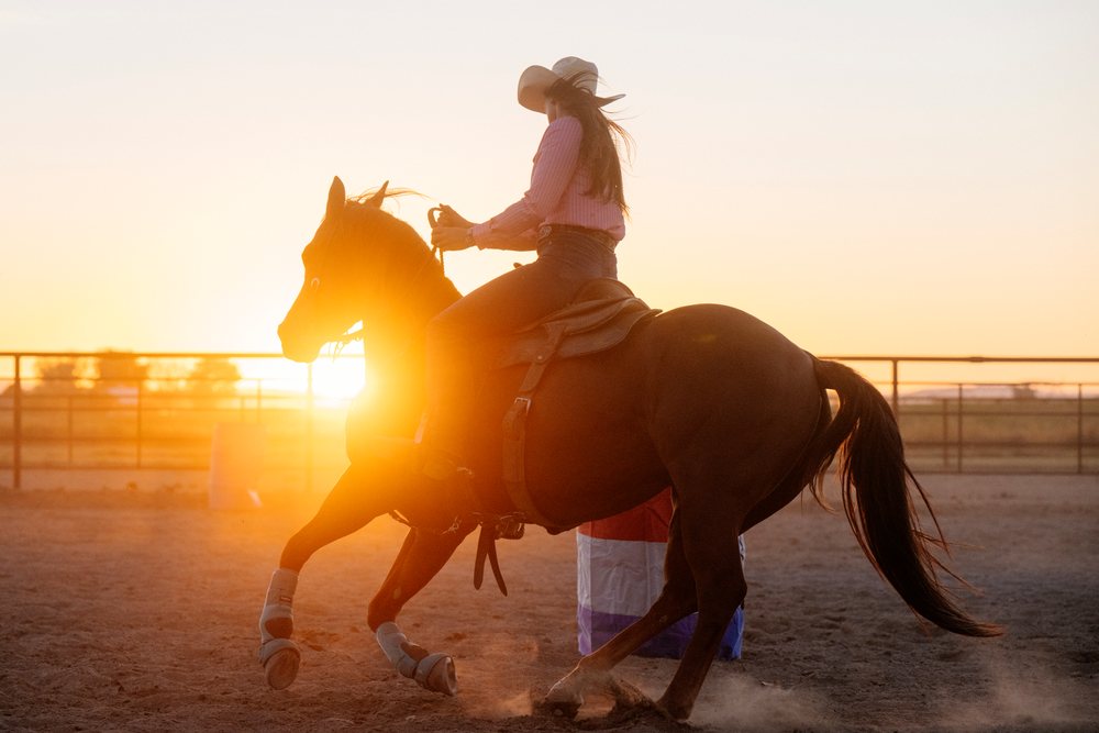 Horseback rider in cowgirl attire sharply turns a horse in a dusty outdoor arena at sunset, Southwest USA.