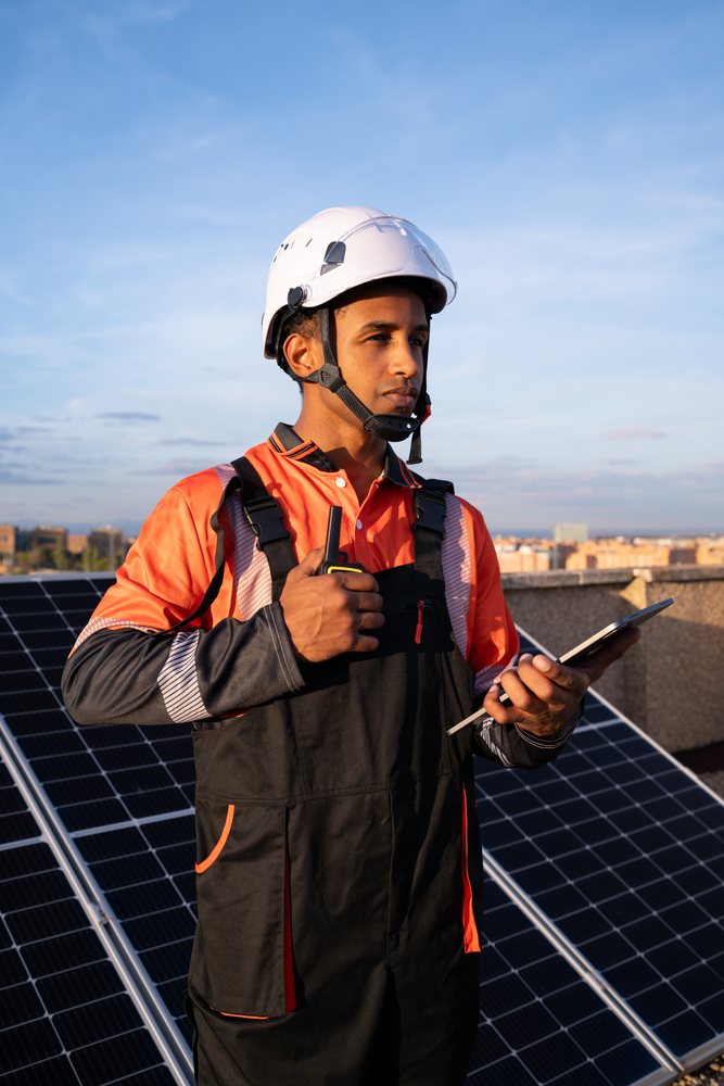 Solar energy technician in a white helmet and work uniform uses a walkie talkie and tablet while standing by rooftop solar panels under a blue sky, Madrid, Spain.