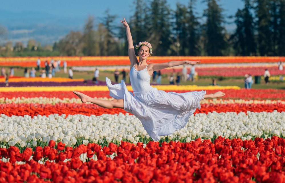 Female Ballet dancer wearing a white skirt and flower crown leaps gracefully in a colorful tulip field during a spring outdoor event, USA.