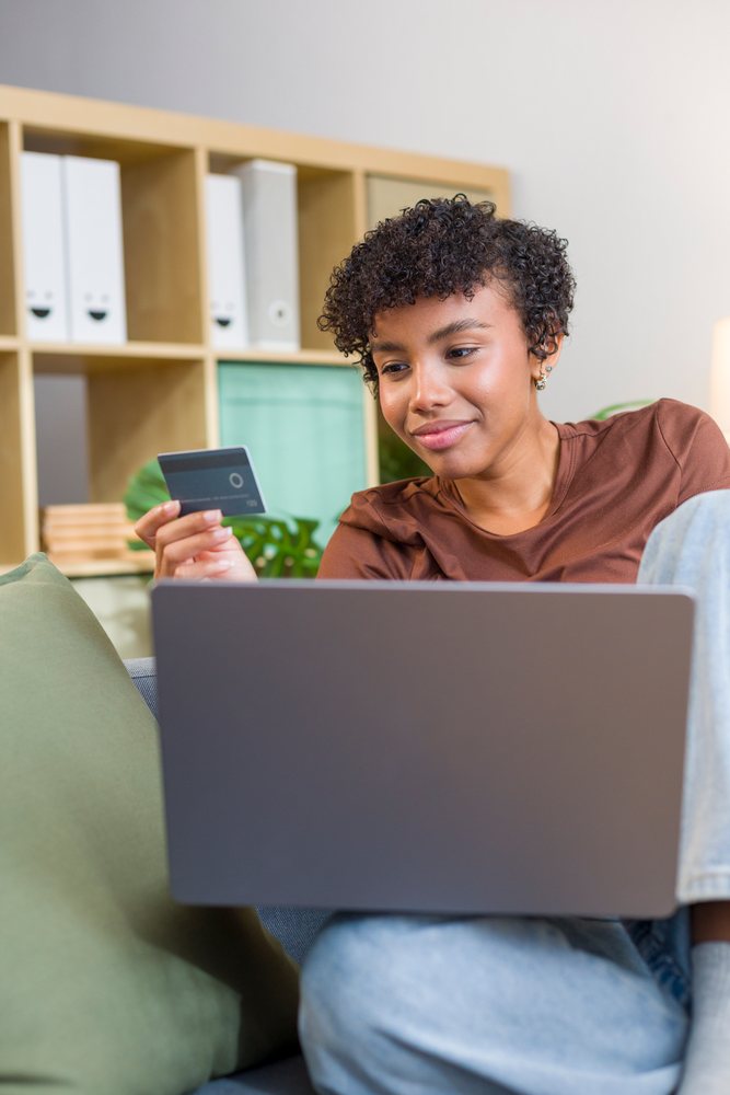 Young woman sitting on a sofa at home using a laptop and holding a credit card with bookshelf and green pillow visible.