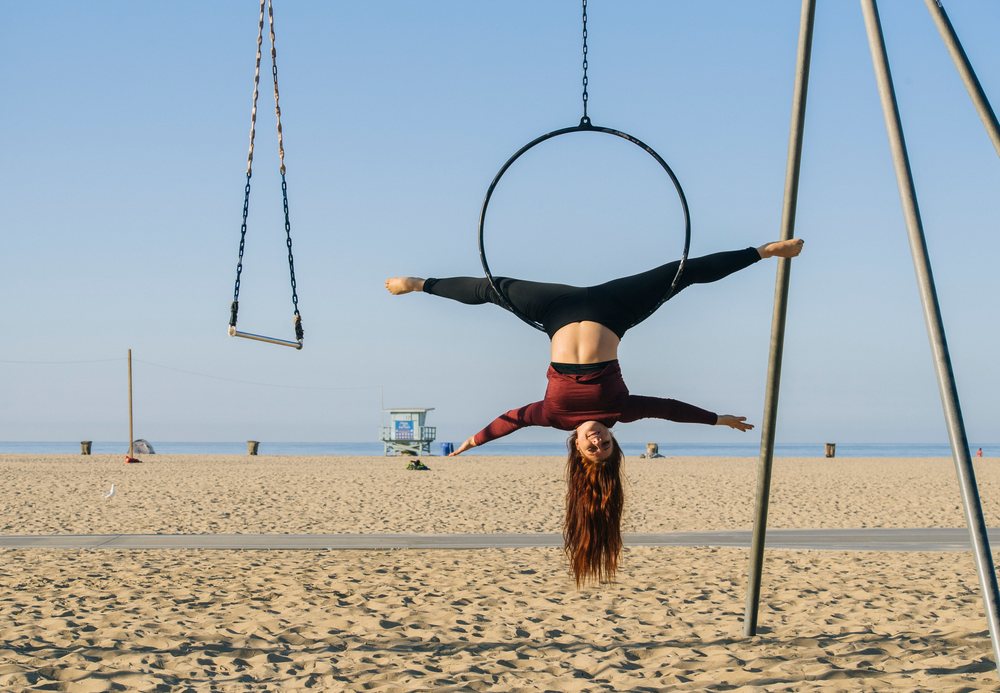 Woman performing a split on a suspended aerial hoop on a sandy California beach near a lifeguard tower under blue sky, USA.