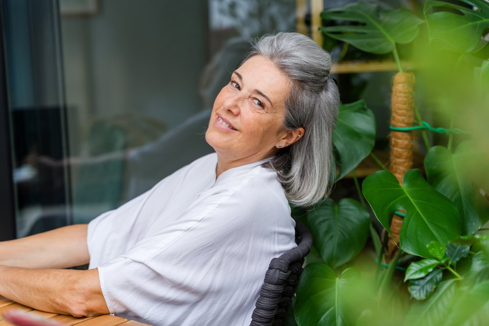 Smiling mature woman with gray hair sitting indoors next to a large leafy plant, Spain.
