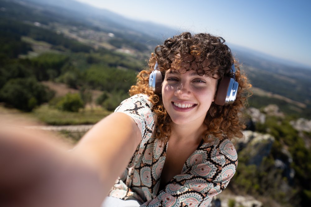 Woman with curly hair wearing headphones takes a selfie atop a mountain overlooking a scenic landscape, Pico Sacro, Galicia, Spain.