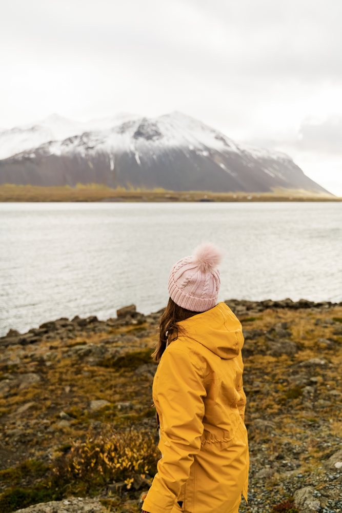 A woman wearing a yellow jacket and pink knit hat looks out over a lake toward a snowy mountain, Iceland.
