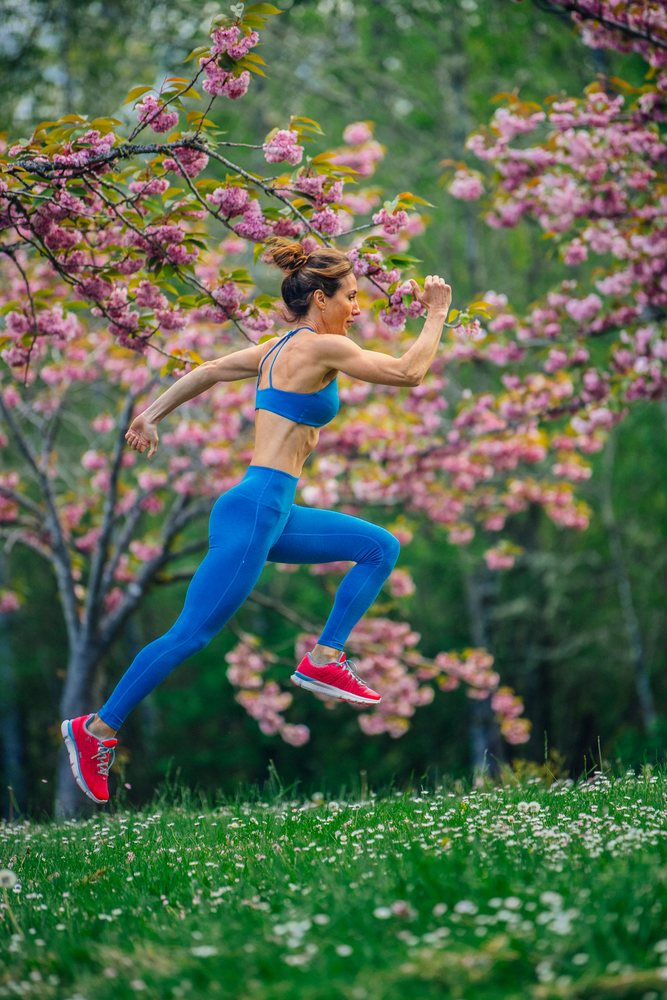 Woman in blue athletic wear jumps energetically outdoors on green grass with wildflowers under blooming pink cherry blossom trees in spring, Calisthenics, USA.