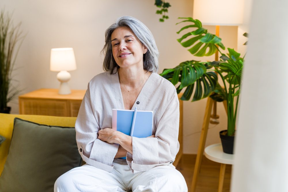 Mature woman sits on a yellow sofa indoors holding a notebook with a potted plant and lamp on a wooden table nearby, Spain.