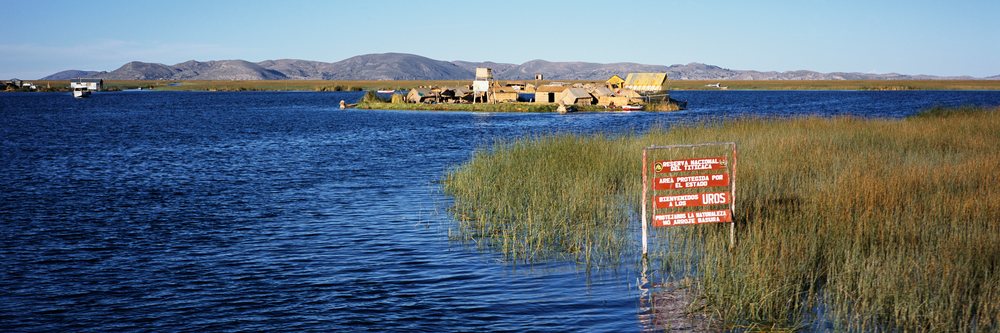 Reed-covered floating islands with wooden and thatched homes lie on Lake Titicaca, backed by mountain ranges under clear blue sky, Uros Floating Islands Lake Titicaca, Peru.