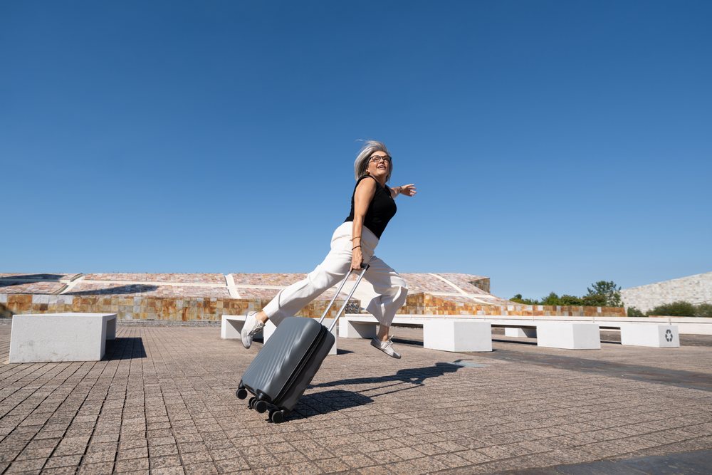 Gray-haired woman wearing glasses jumps while holding a suitcase on a sunny paved plaza in Spain under a clear blue sky, businesswomen.