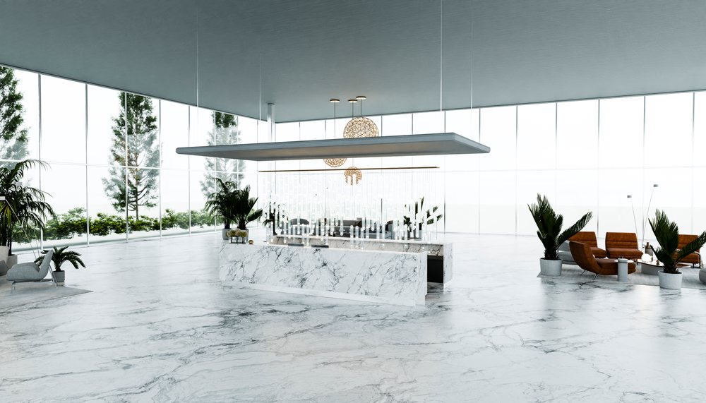 Modern lobby featuring a marble floor, floor-to-ceiling glass windows, reception desk, lounge seating, and multiple green plants under spherical pendant lights.