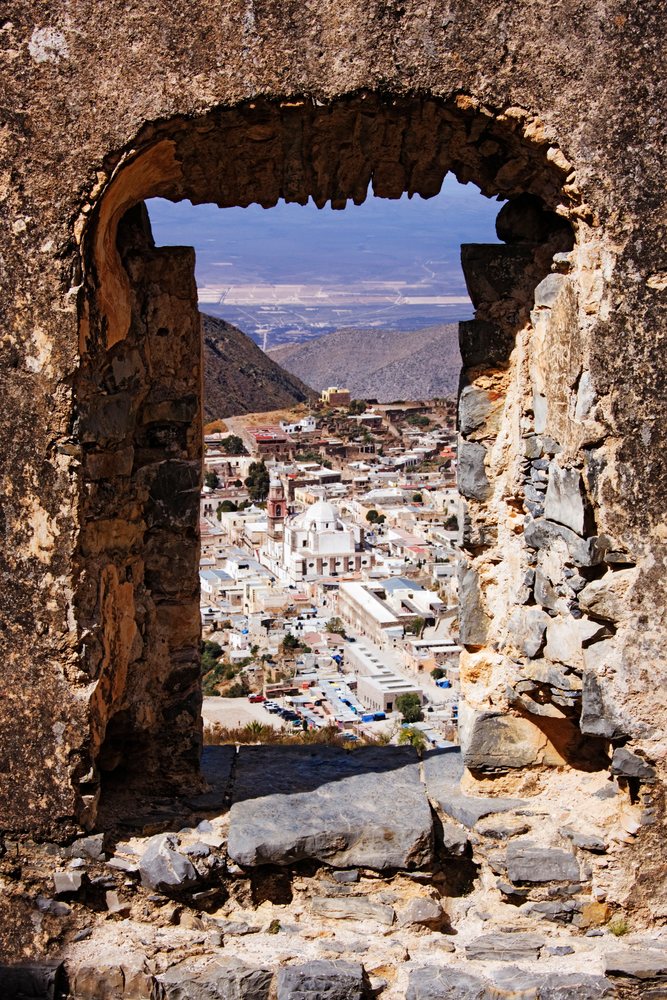 View of Real de Catorce village through a stone archway in ruined walls with surrounding mountains and desert landscape, San Luis Potosi, Mexico.