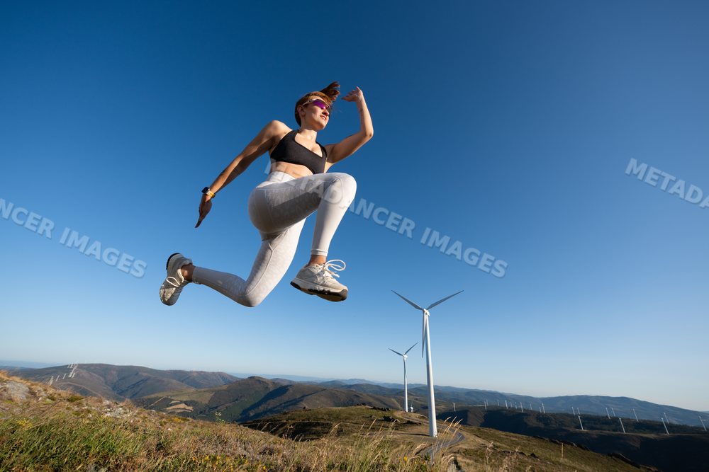 A white woman jumps on a hilltop near wind turbines in Galicia, Spain