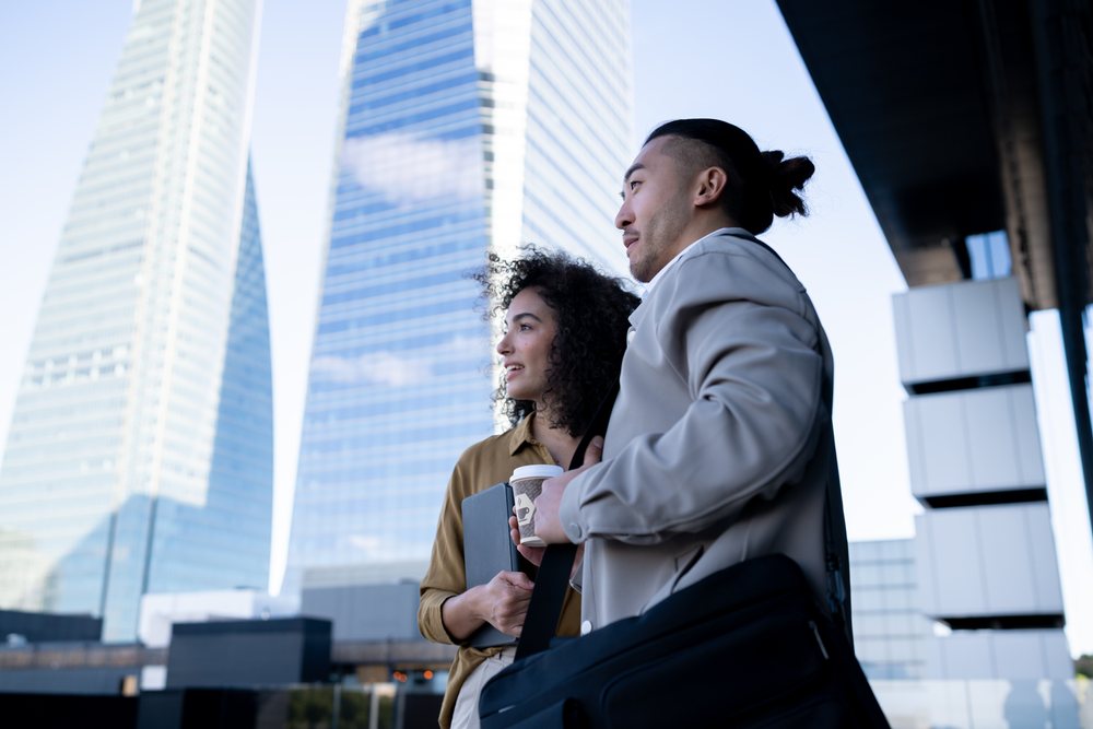 Businessman and businesswoman stand outdoors holding coffee and tablet near modern office buildings in a daytime urban cityscape, Madrid, Spain.
