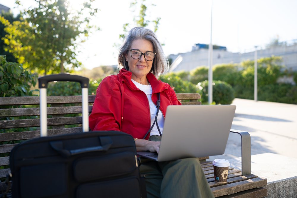 Woman sitting on a bench outdoors, working on a laptop with a black suitcase and a coffee cup nearby, Spain.