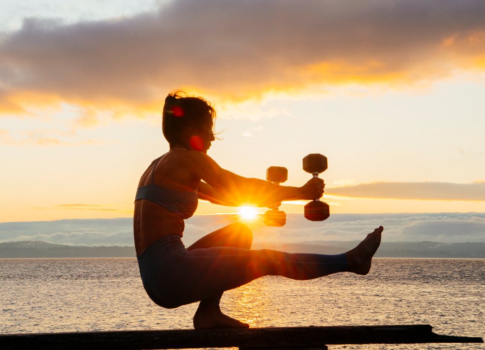 Barefoot woman holding dumbbells performs a balancing squat pose outdoors by a lake at sunset, WA, USA.