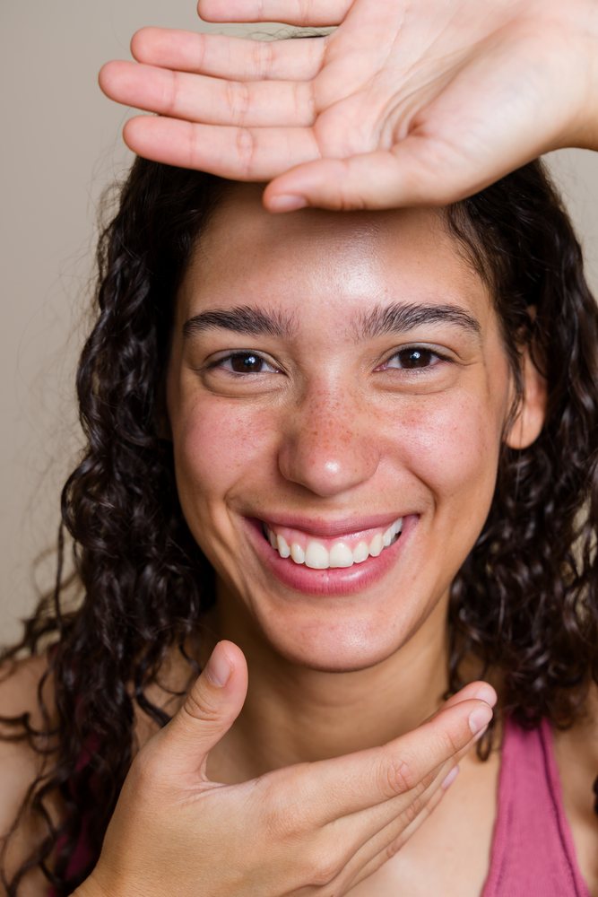 Woman with curly hair, freckles, and brown eyes smiling with hands near her face in a beauty shoot, Spain.