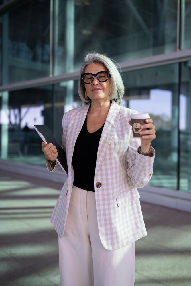 Middle-aged businesswoman with gray hair wearing glasses and a plaid blazer stands outside a modern glass office building holding a tablet and coffee cup, Spain, Madrid.